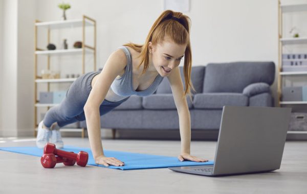 Online sport exercises at home. Workout at home. Redhead girl doing exercises at home. Young athletic woman push-ups is watching a video in a laptop in a living room.